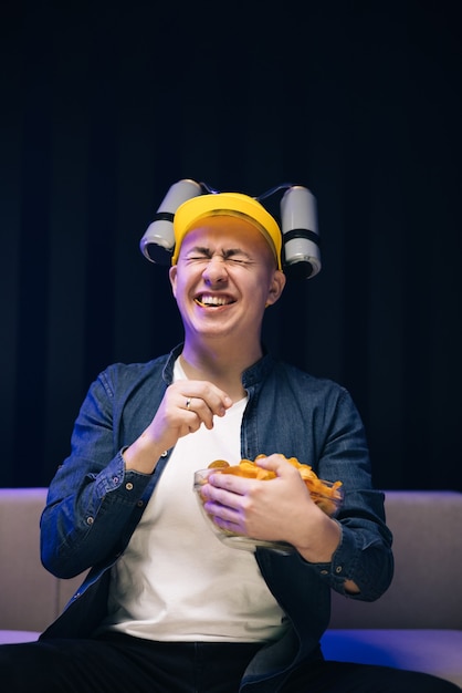 Handsome man with beer helmet on the head watching tv and eating potato chips on the couch at home