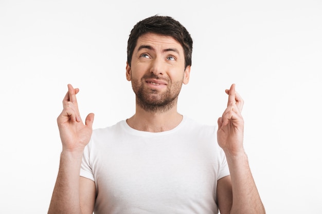 handsome man with bristle in casual t-shirt keeping fingers crossed and making a wish isolated over white wall