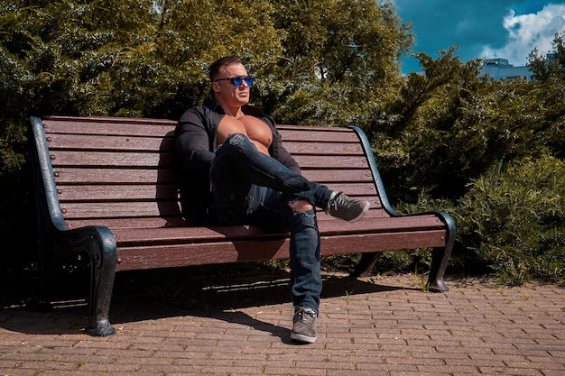 handsome man with glasses wearing a black shirt and jeans sits on a bench in the park