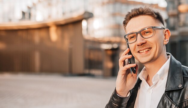 Handsome man with glasses with a smartphone on the street of a big city. Businessman talking on the phone on urban background