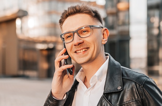 Handsome man with glasses with a smartphone on the street of a big city. Businessman talking on the phone on urban background