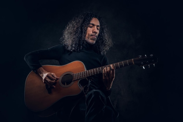 Handsome man with long hair is playing acoustic guitar at dark studio.