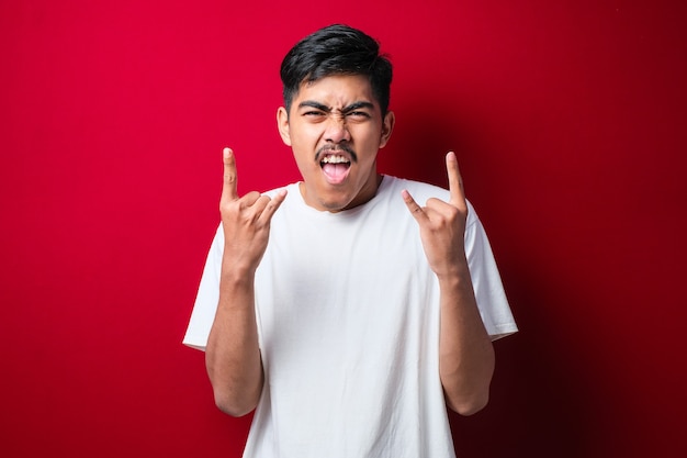 Handsome man with mustache wearing white tshirt over red background shouting with crazy expression doing rock symbol with hands up. Music star. Heavy concept.