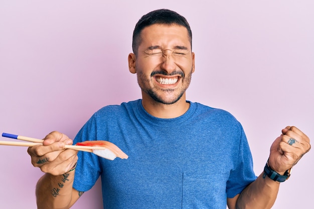 Handsome man with tattoos eating sushi using chopsticks screaming proud, celebrating victory and success very excited with raised arm