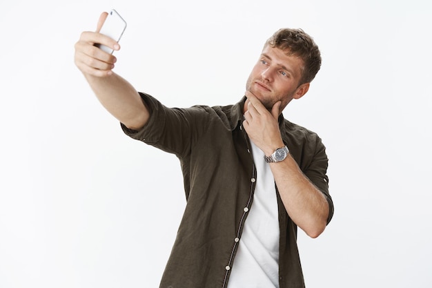 Handsome and masculine macho man making cool pose touching bristle on jaw and taking selfie with sexy smile