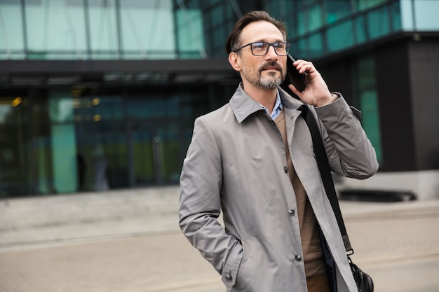 handsome mature businessman in eyeglasses talking on cellphone while standing near office building in urban area