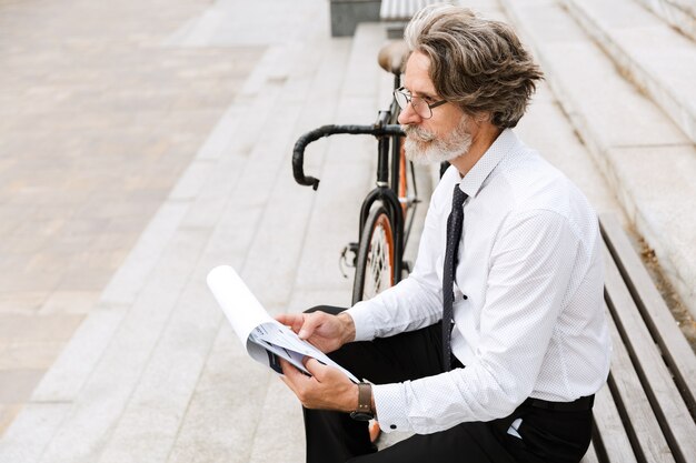 Handsome mature businessman in formalwear sitting on a bench outdoors, analyzing documents while holding a clipboard