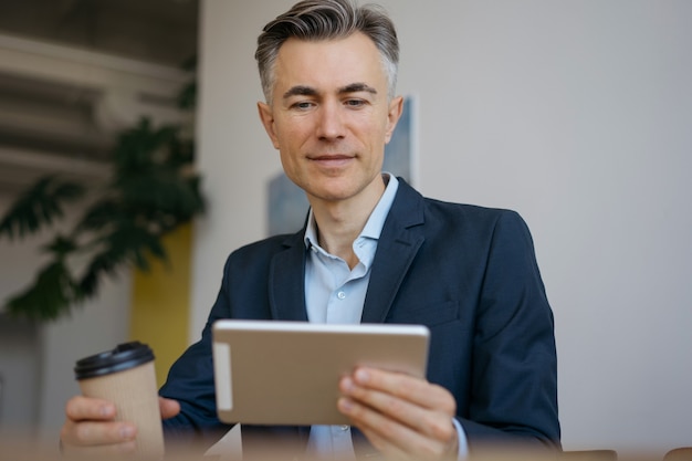 Handsome mature businessman using digital tablet in office. Successful freelancer holding cup of coffee working from home