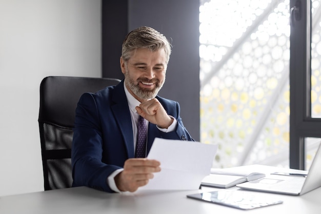 Handsome Mature Male Entrepreneur Reading Papers While Sitting At Desk In Office