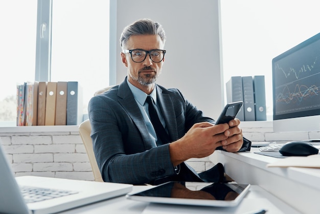 Handsome mature man in formalwear holding smart phone while sitting in office