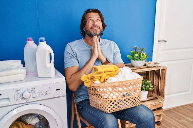 Handsome middle age man waiting for laundry praying with hands together asking for forgiveness smiling confident