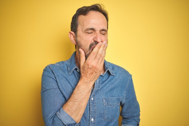 Handsome middle age senior man with grey hair over isolated yellow background bored yawning tired covering mouth with hand Restless and sleepiness