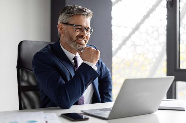 Handsome middle aged businessman in suit sitting at workplace in modern office