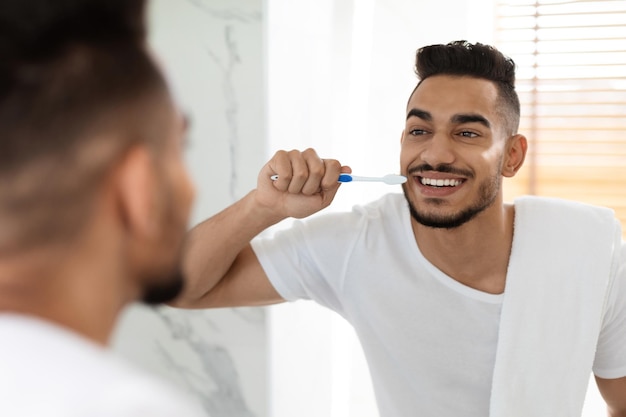 Handsome millennial arab guy brushing his teeth with toothbrush in bathroom