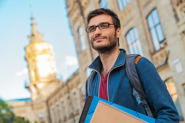 Handsome mixed race male student in eyeglasses with books outside near campus