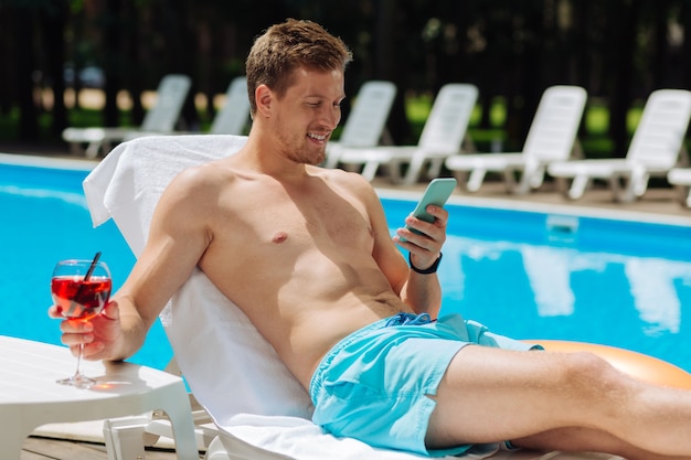 Handsome muscle man reading message on his blue smart phone while getting suntan