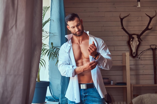 A handsome muscular Caucasian male dresses a white shirt near the window in the morning.