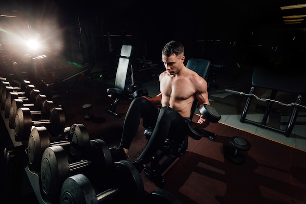 Handsome muscular male exercising with heavy dumbbells while standing in dark modern gym