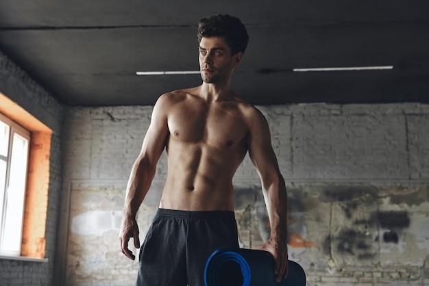 Handsome muscular man carrying exercise mat while standing in gym