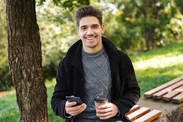 handsome optimistic young happy man in casual clothing walking outdoors in green park using mobile phone drinking coffee.