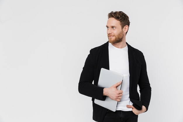 handsome pleased man in jacket smiling and holding laptop isolated over white wall