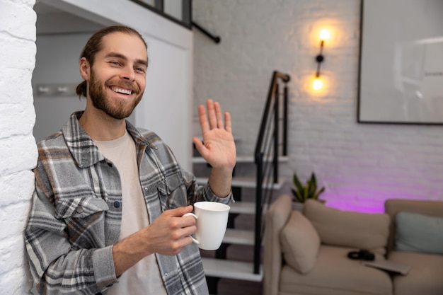 Handsome positive man holding cup of fresh coffee or tea in hands enjoying free time with hot beverage waving hand saying hello