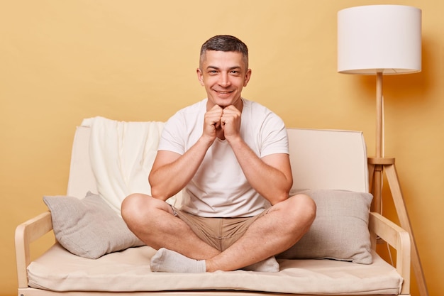 Handsome positive optimistic man wearing white Tshirt and shorts sitting on sofa against beige background looking at cmaera with delighited expression happy face