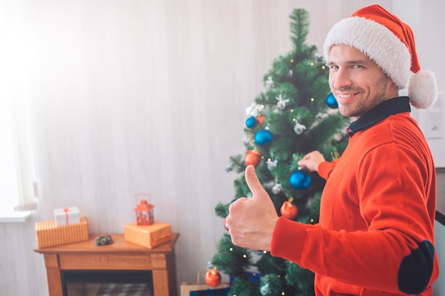 Handsome and positive young man stand and shows big thumb up with one hand and put toy on Christmas tree with another one