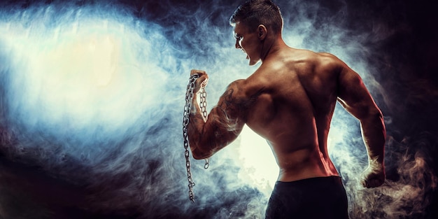 Handsome power athletic man bodybuilder doing exercises with chain fitness muscular body on dark bac