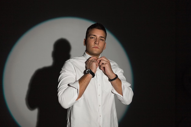 Handsome professional man in a fashion white shirt buttoning up a button in a dark background in a studio with a circular light at a party