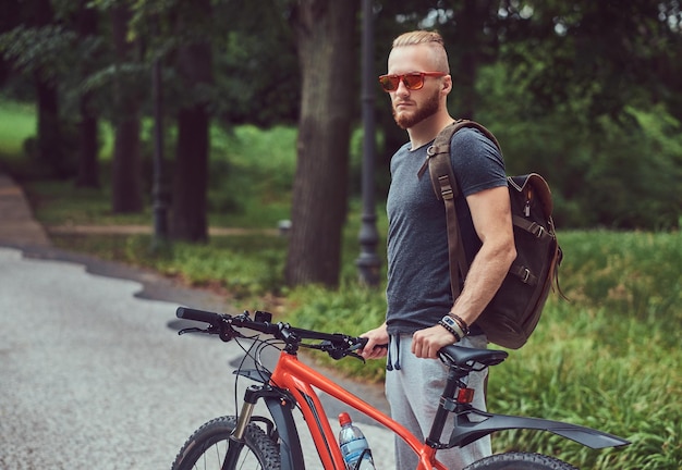 Handsome redhead male with a stylish haircut and beard dressed in sportswear and sunglasses walks in the park with a bicycle and backpack.