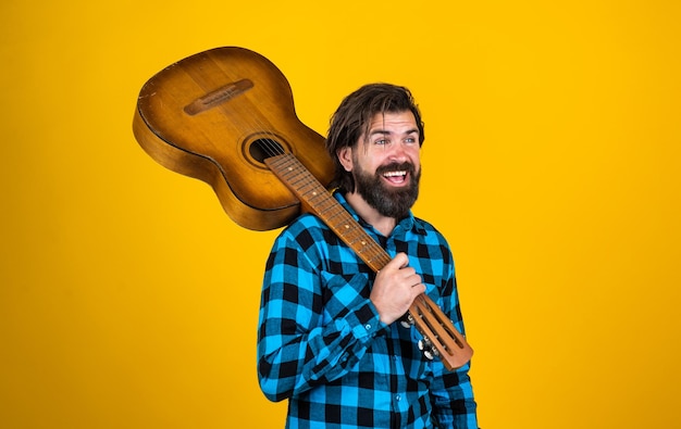 Handsome rock star wearing checkered shirt posing while holding his acoustic guitar and standing on yellow background music