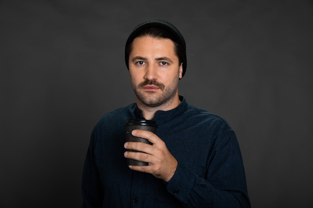 Handsome sad unshaven guy in knitted cap posing with disposable cup of coffee