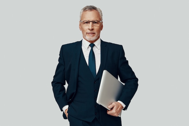 Handsome senior man in full suit carrying laptop and looking at camera while standing against grey background