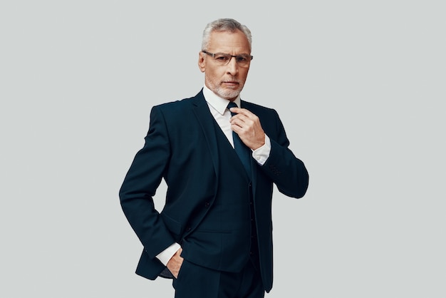 Handsome senior man in full suit looking at camera and adjusting his tie while standing against grey background