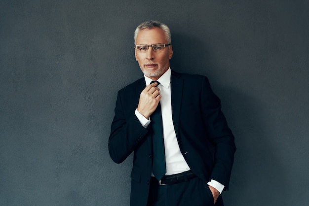 Handsome senior man in full suit looking at camera and adjusting tie while standing against grey background