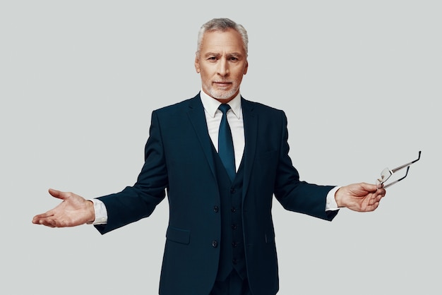 Handsome senior man in full suit looking at camera and keeping arms outstretched while standing against grey background