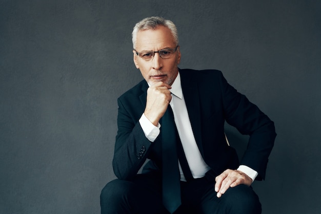 Handsome senior man in full suit looking at camera and keeping hand on chin while sitting against grey background