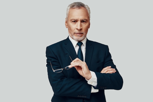 Handsome senior man in full suit looking at camera and keeping hand on chin while standing against grey background