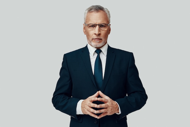 Handsome senior man in full suit looking at camera and keeping hands clasped while standing against grey background