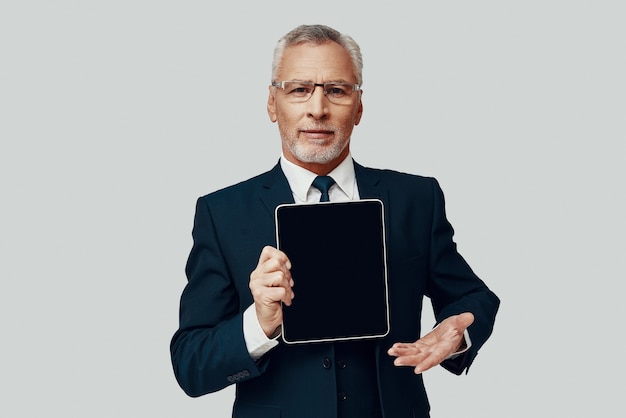 Handsome senior man in full suit pointing copy space on digital tablet and smiling while standing against grey background