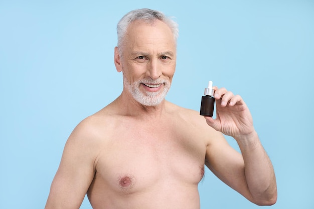 Handsome senior man showing a jar with anti aging serum smiling over blue background