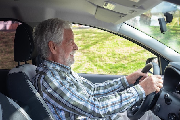 Handsome senior man with checkered shirt sitting inside his car smiles while looking ahead Smiling bearded elderly grandfather driving the auto with hands on steering wheel