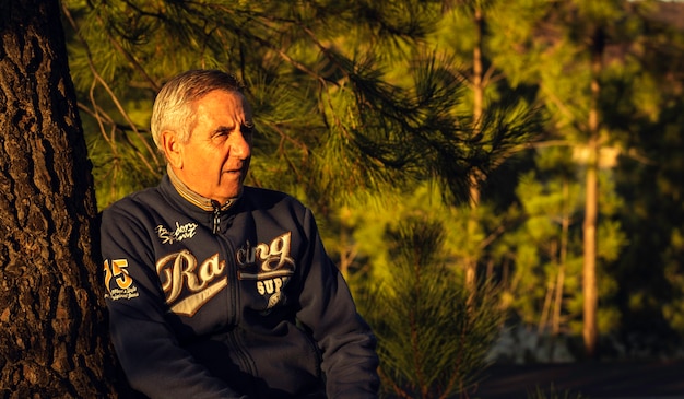 Handsome senior man with sportwear resting on a tree at the woodland  and looking afar