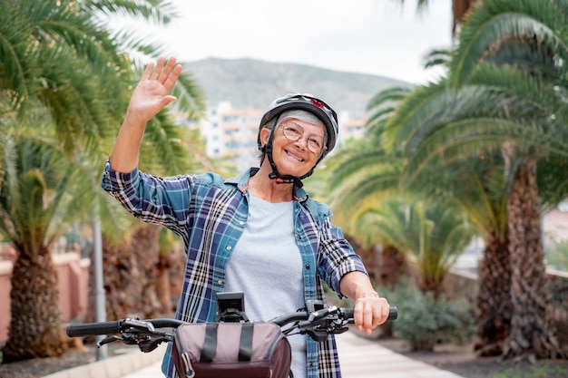 Handsome senior woman with helmet riding electric bicycle in park waving hand enjoying healthy life