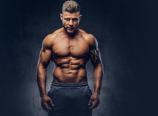 A handsome shirtless bodybuilder with stylish haircut and beard, with tattoo on his arm, posing in a studio. Isolated on a dark background.