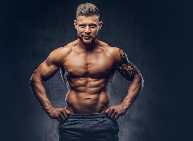 A handsome shirtless bodybuilder with stylish haircut and beard, with tattoo on his arm, posing in a studio. Isolated on a dark background.