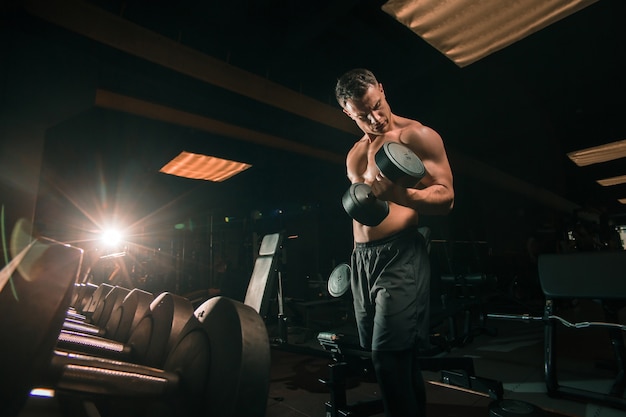 Handsome shirtless guy lifting heavy dumbbells while sitting on bench in dark gym