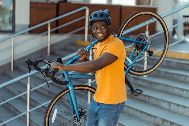 handsome smiling African American man wearing helmet holding his bicycle walking on urban street