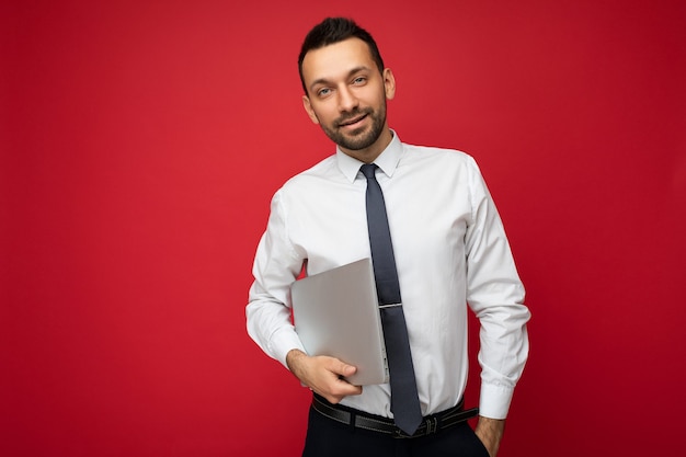 Handsome smiling brunet man holding laptop computer  in white shirt and tie on isolated red wall.